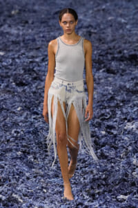 Model walking on a runway in a light gray sleeveless top and shredded denim fringe shorts, mid-stride on a dark blue textured runway.