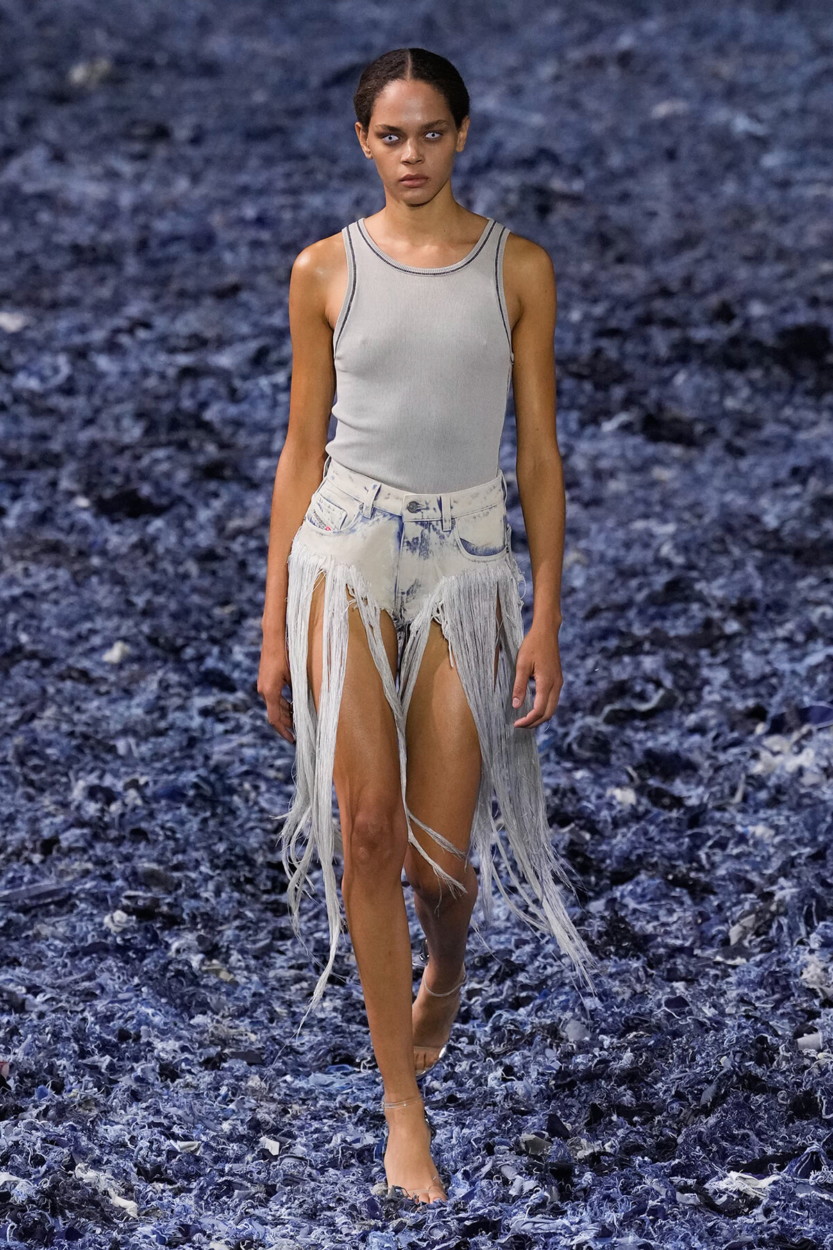 Model walking on a runway in a light gray sleeveless top and shredded denim fringe shorts, mid-stride on a dark blue textured runway.