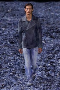 Male model walking on a blue-toned runway in a dark denim jacket and light-wash jeans.
