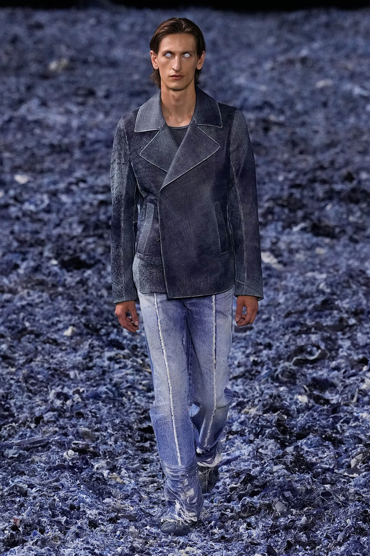 Male model walking on a blue-toned runway in a dark denim jacket and light-wash jeans.