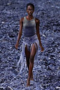 Model walking a fashion runway in a gray sleeveless top and torn denim shorts with long fringe, against a blue-textured backdrop.