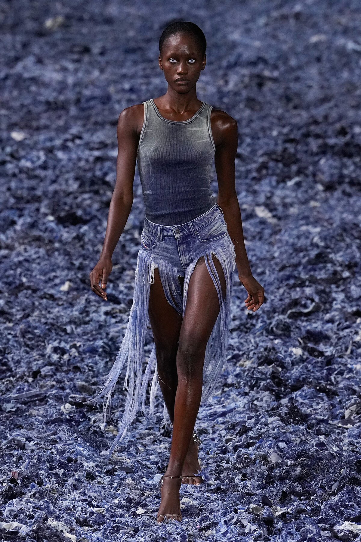 Model walking a fashion runway in a gray sleeveless top and torn denim shorts with long fringe, against a blue-textured backdrop.