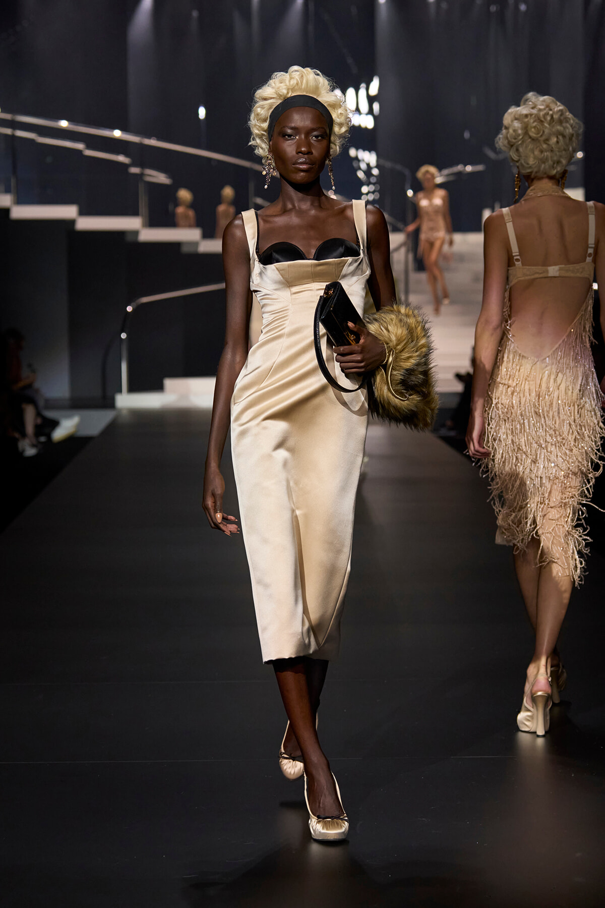 Model walks a runway in a cream satin midi dress with a black bustier, holding a black clutch and fur stole.