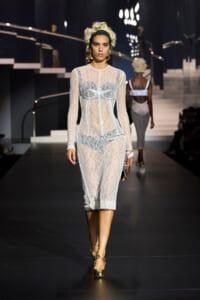 Model walks a fashion show wearing a sheer white lace bodysuit with a corseted bodice, headband, and gold jewelry on a dark runway.