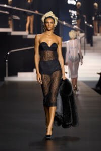 Model walking the runway in a black strapless lace dress with sheer panel at the waist, holding a black fur coat.