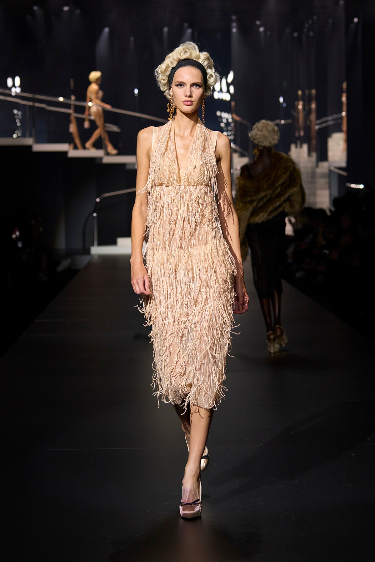 Model walks the runway in a cream/beige fringed dress with a halter neckline, vintage curls, and gold earrings.