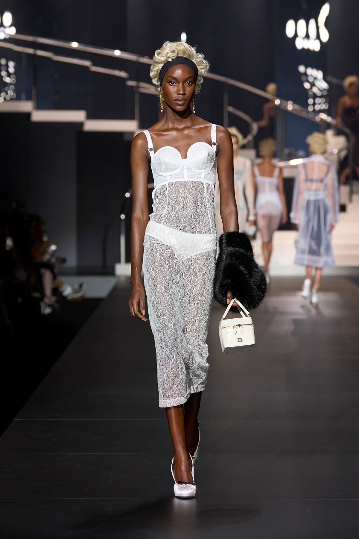 Black model walking a fashion runway in a white lace lingerie-style dress with a corset top, headband, and white heels, carrying a small white handbag and black fur stole