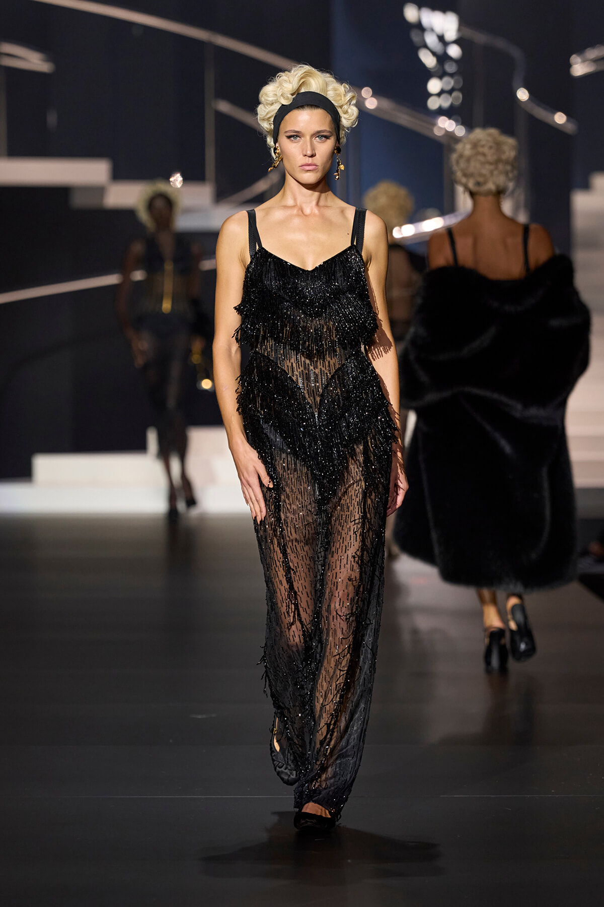 Model walking the runway in a sheer black beaded fringe gown with a blonde updo and headband.