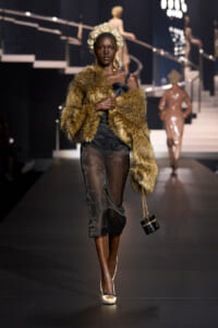 Model on a runway wearing a brown fur stole over a sheer black dress, with a white curled wig and a small black handbag with a gold chain side-slung.