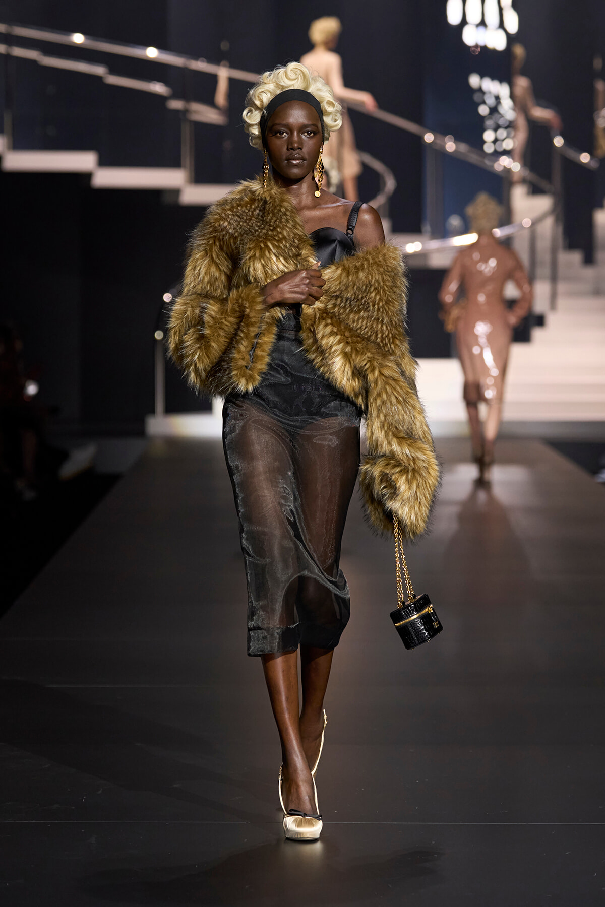 Model on a runway wearing a brown fur stole over a sheer black dress, with a white curled wig and a small black handbag with a gold chain side-slung.