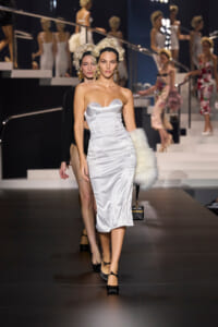Model walks the runway in a white strapless satin midi dress with a fur stole, gold chandelier earrings, and black heels.