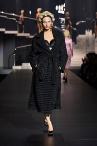 Female model on a fashion runway wearing a long black textured wrap coat with a belt, revealing a black bustier beneath.