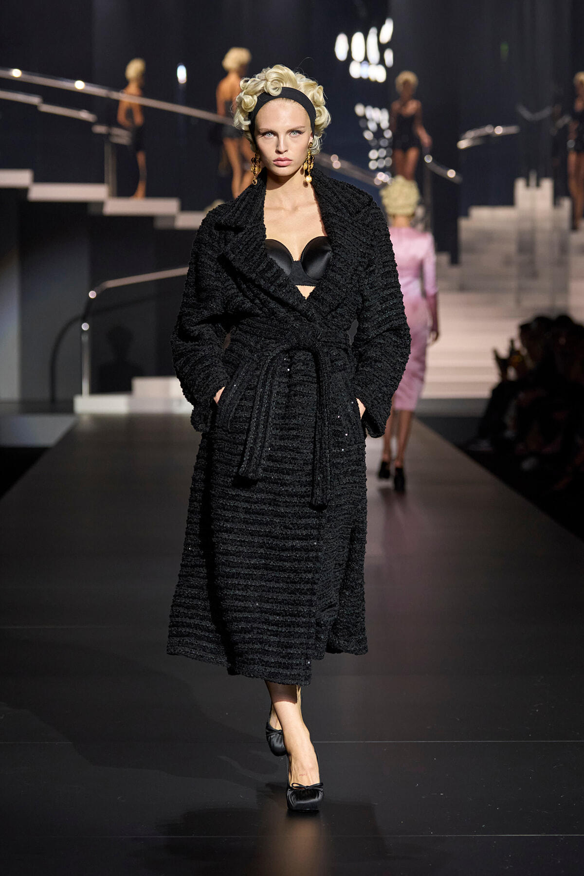 Female model on a fashion runway wearing a long black textured wrap coat with a belt, revealing a black bustier beneath.