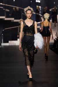 Model walks the runway in a black beaded fringed gown, carrying a white fur stole and a small round clutch.