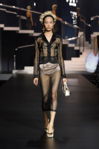 Model walking a fashion runway in a sheer black lace suit with a gold underlayer, black headband, gold earrings, holding a small white handbag.