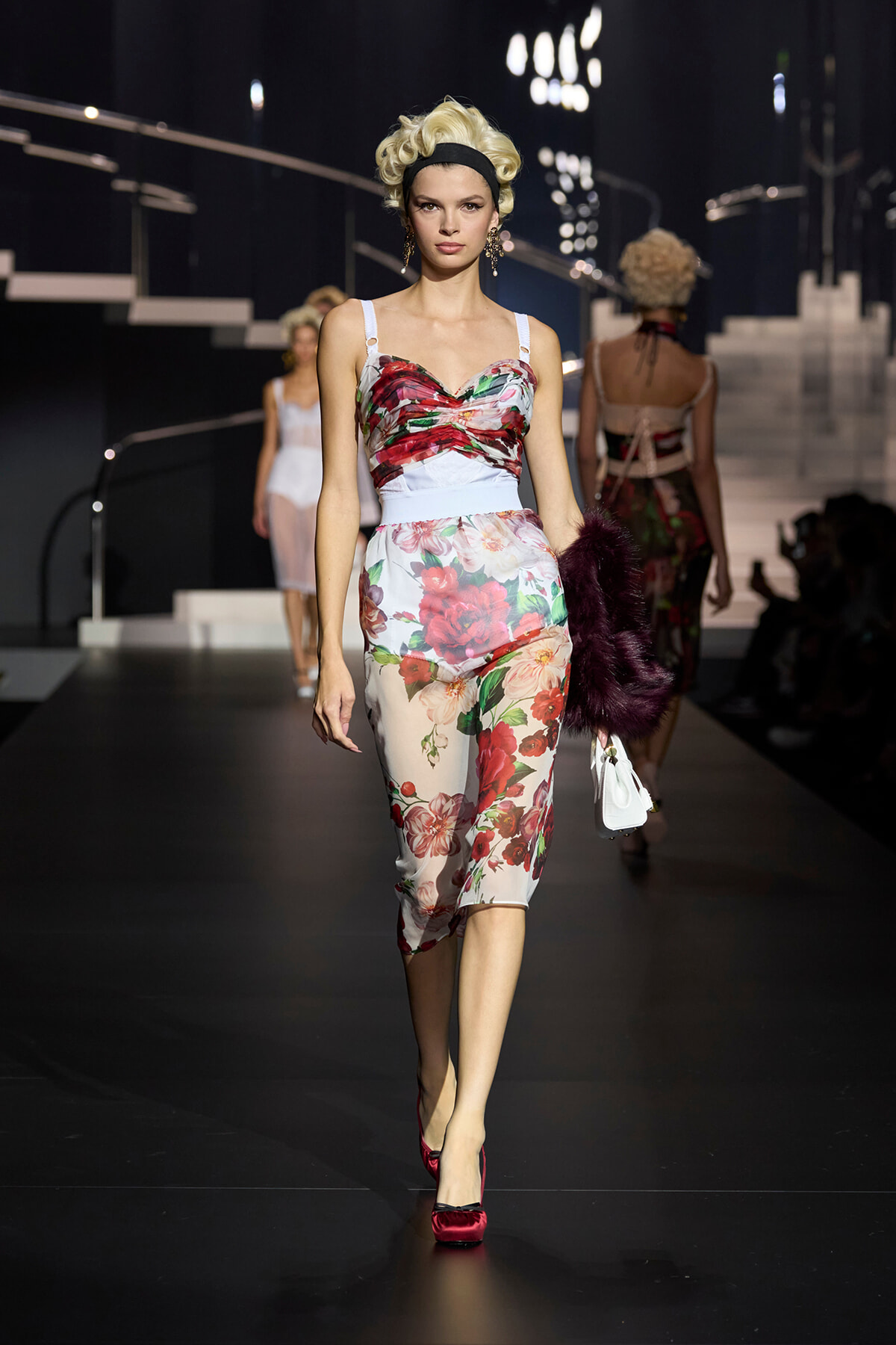 Model walks the runway in a white floral two-piece dress with red and pink blooms and a light blue waistband, red heels, and a black headband.