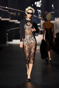 Runway model walking in a sheer black lace midi dress with a cropped black jacket, black heels, oversized sunglasses, and a black clutch.