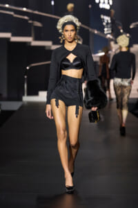 Model walks a fashion runway in a black lingerie-inspired outfit: cropped cardigan over a black bra, high-waisted shorts with garters, platinum wig, large cross earrings, and black fur clutch.