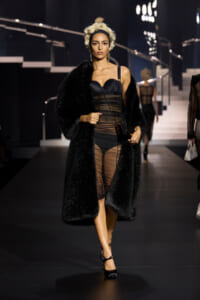Model walks a runway in a black sheer bodysuit with sheer panels, complemented by a long black fur coat and black heels.
