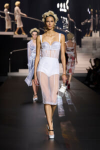 Model walking the runway in a white sheer corset-style dress with a sheer skirt, carrying a white handbag.