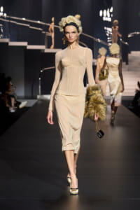 Model walking a beige, form-fitting dress with corset seams on a dark fashion runway, holding a fur stole and small purse.