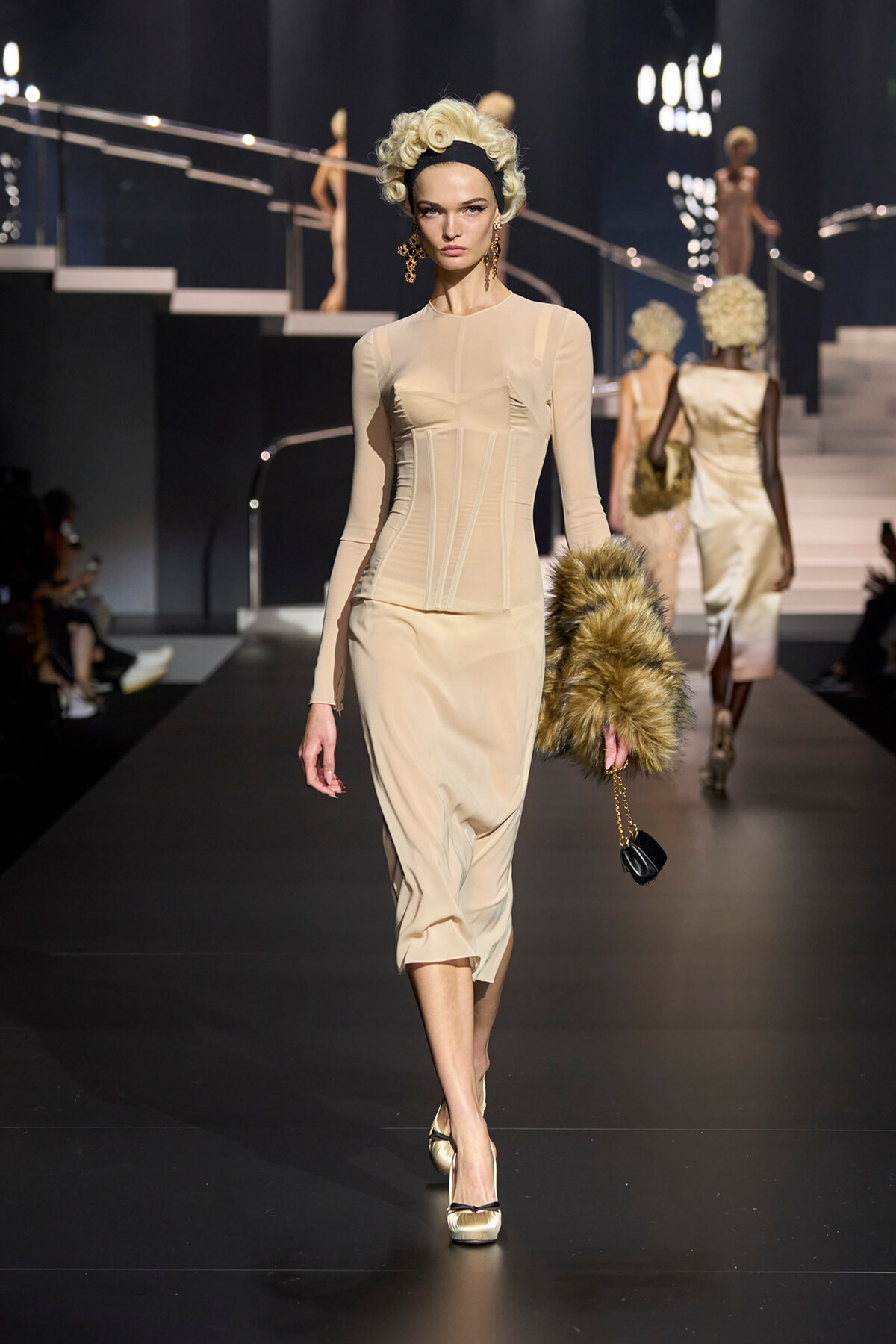 Model walking a beige, form-fitting dress with corset seams on a dark fashion runway, holding a fur stole and small purse.
