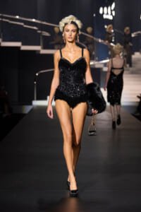 Model walks the runway in a black beaded fringe bodysuit with thin straps, carrying a small black fur stole and handbag with a gold chain.