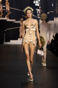 Model walks a fashion show wearing a champagne satin lingerie-inspired set: corset-style top, high-waisted shorts with garter details, and matching heels with a headband.