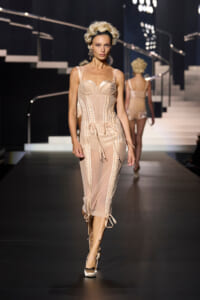 Model walking the runway in a nude-toned corset dress with lace-up panels and sheer inserts on a fashion show catwalk.
