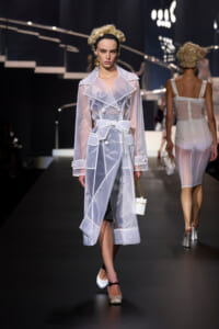 Model walking the runway in a sheer white trench coat with oversized lapels and a wide bow belt, holding a small white handbag and wearing white heels with a black ankle strap.