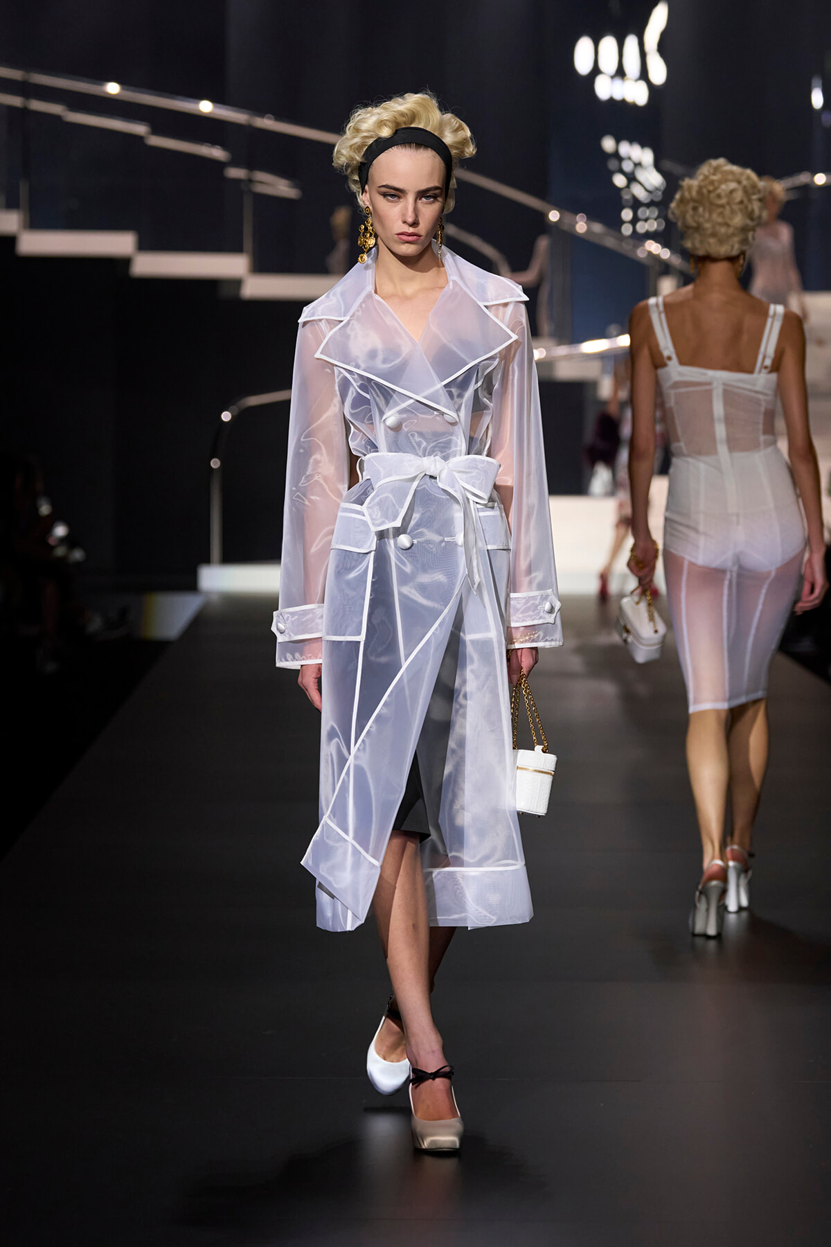 Model walking the runway in a sheer white trench coat with oversized lapels and a wide bow belt, holding a small white handbag and wearing white heels with a black ankle strap.