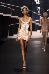 Model walks the runway in a cream corset top and matching high-waisted shorts with garter straps, beige heels, and a vintage curl wig.