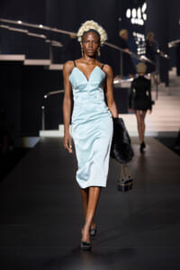 Model walks a runway in a pale blue satin midi dress, wearing a dramatic white wig, black heels, and carrying a black fur accessory with a small black handbag on a chain.