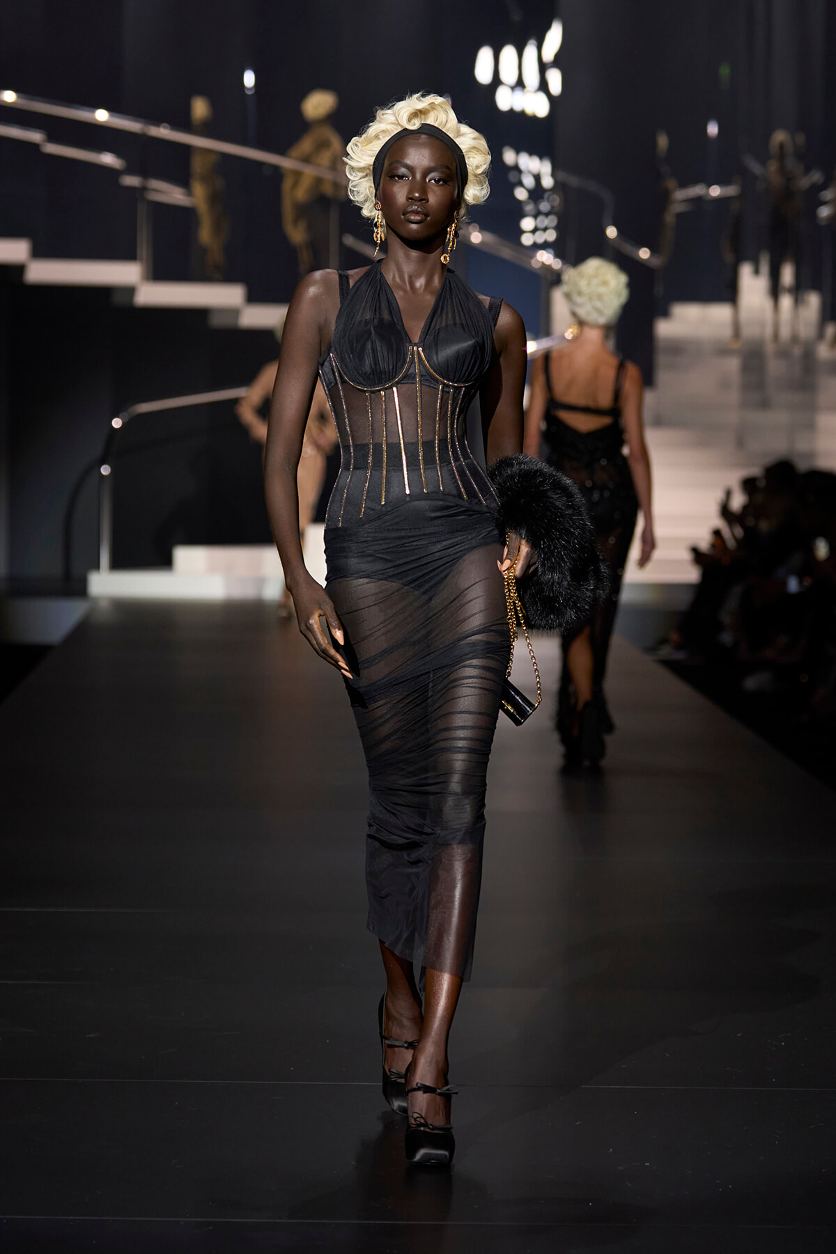 Runway model in a black sheer gown with gold corset detailing and platinum blonde hair walking the fashion show.