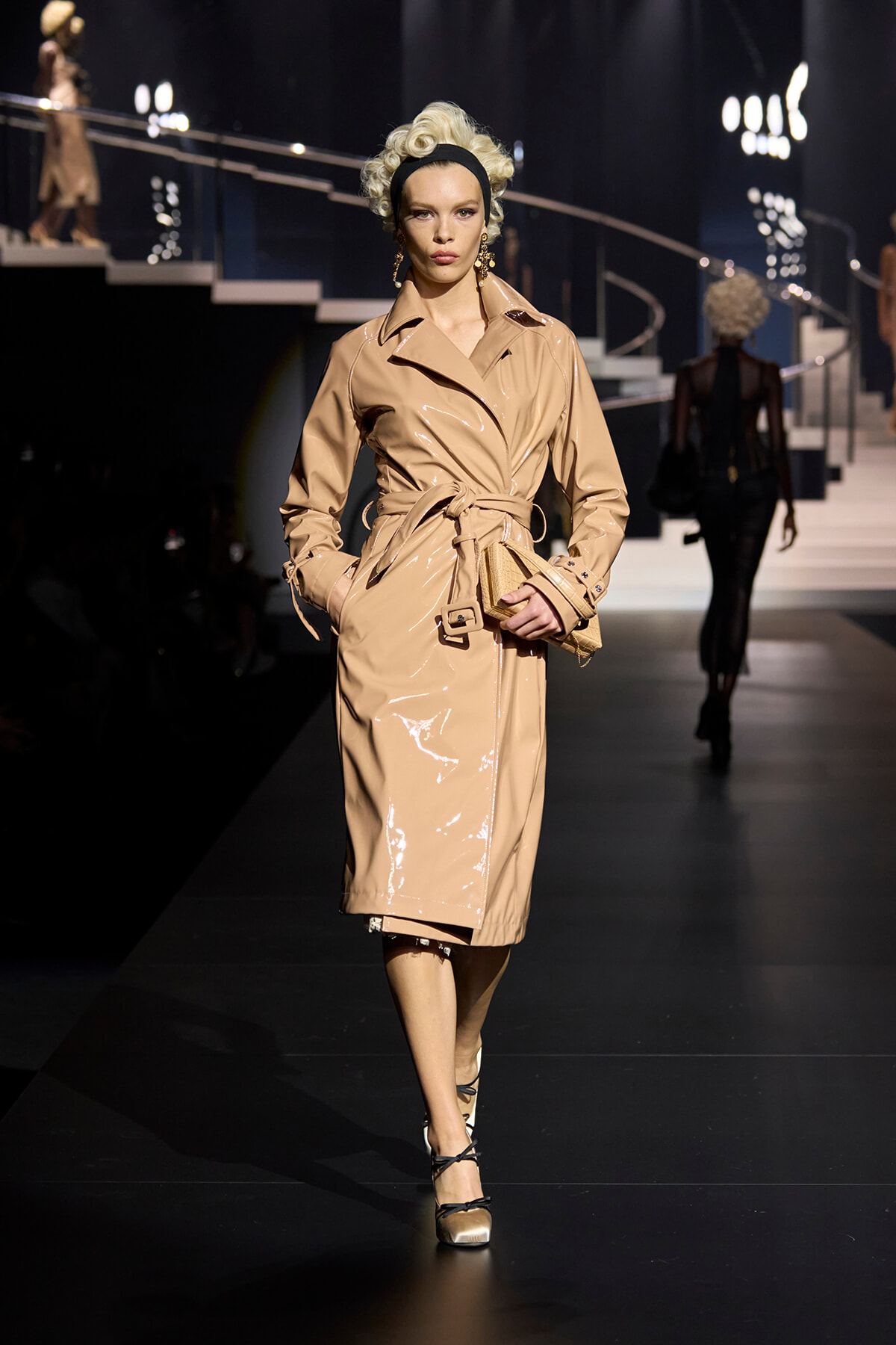 Model walking a fashion runway in a glossy beige trench coat with belt, headband, and gold earrings in a dimly lit venue.” ,