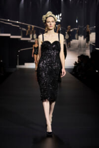 Model walks a backstage-lit runway in a knee-length, black sequin fringed dress with thin straps and vintage hairstyle.