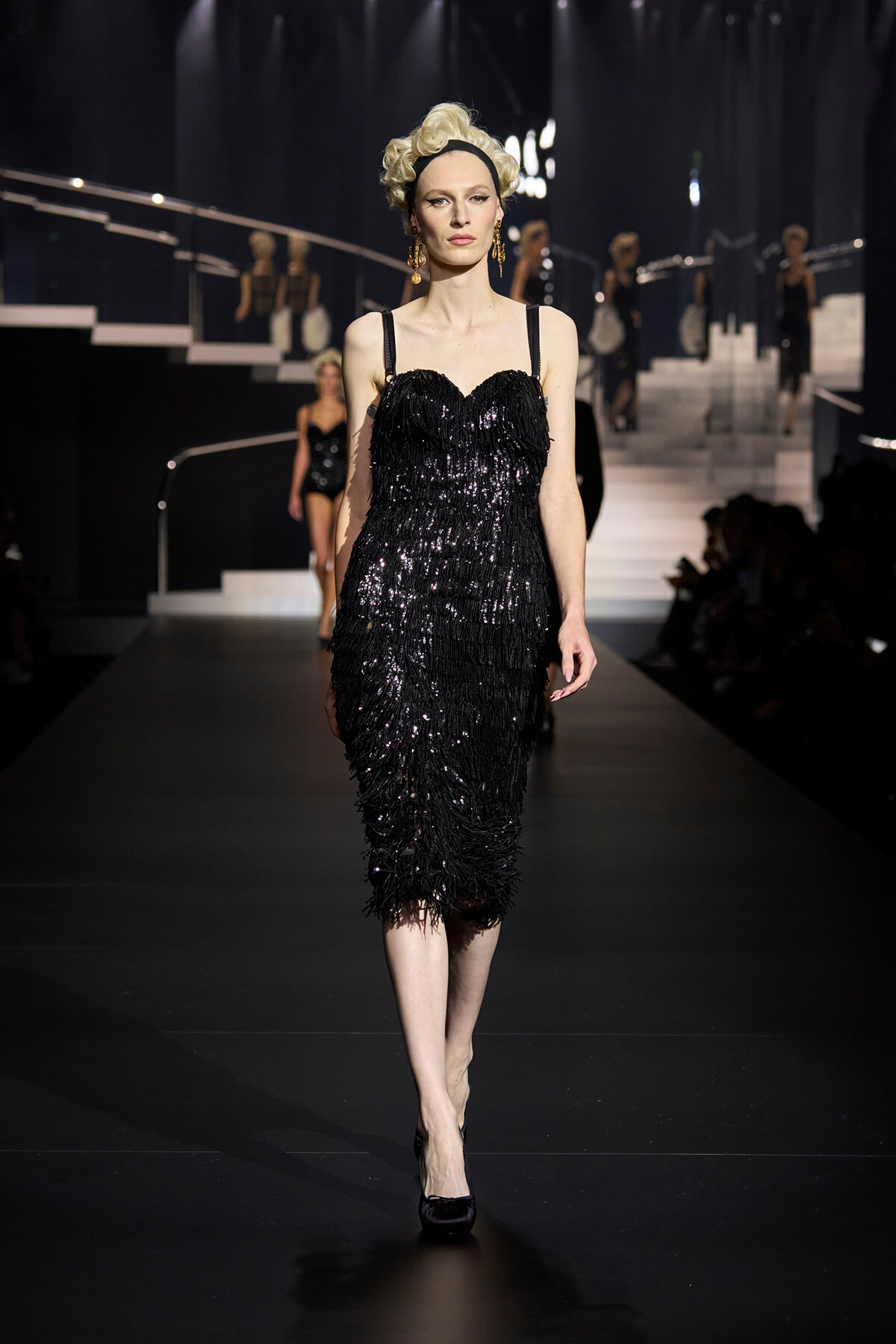 Model walks a backstage-lit runway in a knee-length, black sequin fringed dress with thin straps and vintage hairstyle.