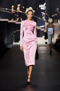 Model walks a pink satin midi dress on a fashion runway, holding a black clutch and wearing black heels with a vintage updo hairstyle.