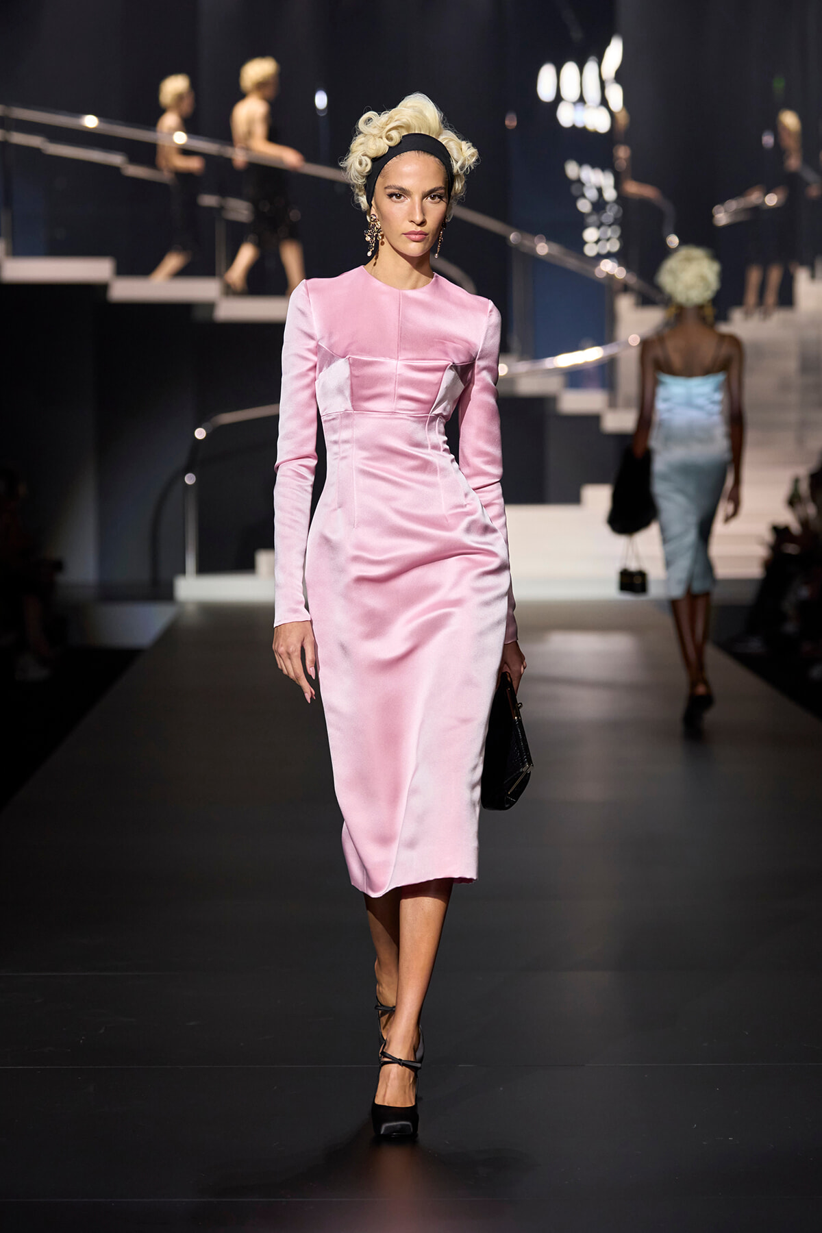 Model walks a pink satin midi dress on a fashion runway, holding a black clutch and wearing black heels with a vintage updo hairstyle.