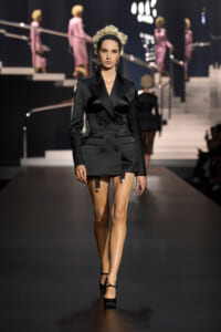 Model walks the runway in a tailored black double-breasted blazer dress with garter straps and black heels.