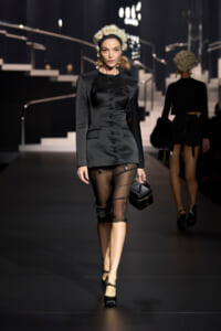 Runway model in a black tailored blazer-dress with front buttons, paired with a sheer black midi skirt and black heels, carrying a small black handbag.