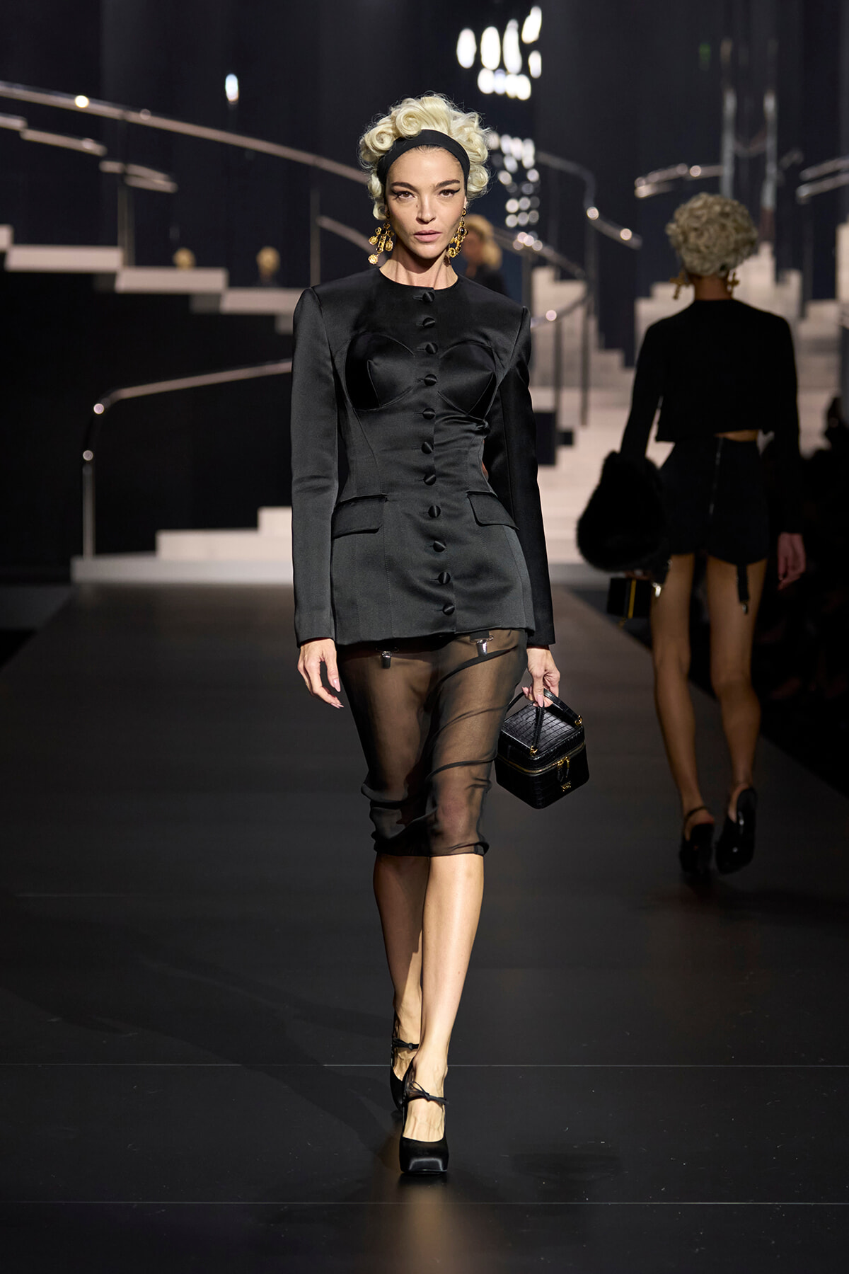 Runway model in a black tailored blazer-dress with front buttons, paired with a sheer black midi skirt and black heels, carrying a small black handbag.