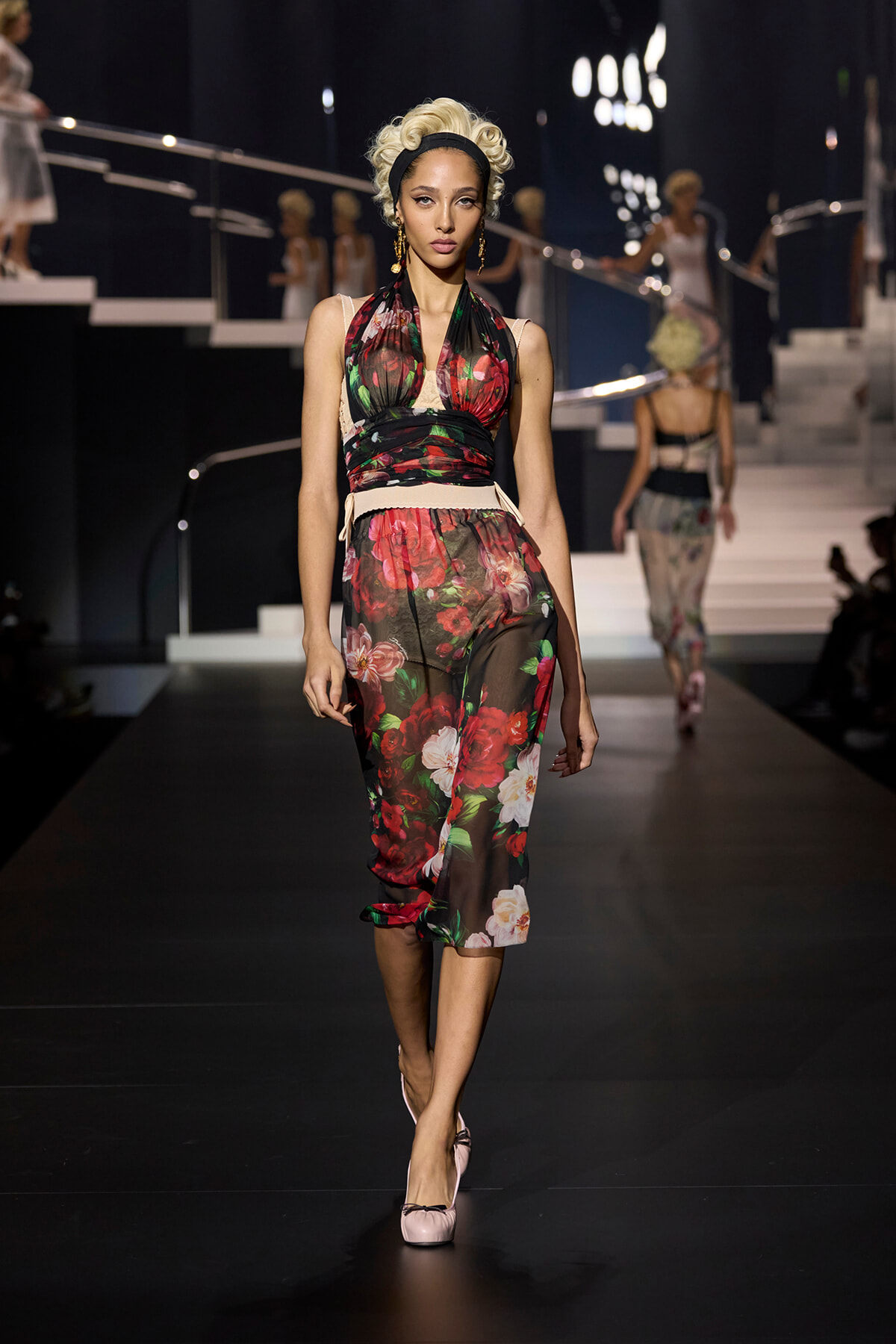 Model walks the runway in a sheer floral jumpsuit with a pale beige belt, pink flats, and a retro headband.
