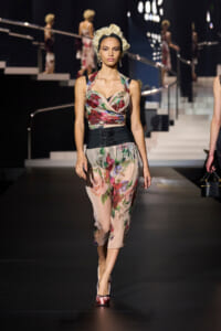 Model struts a floral two-piece ensemble on a runway, featuring a pink-beige bustier top, sheer floral pants, and a black waist cincher, with a vintage curled hairstyle and pink satin heels.