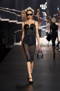 Model walking a fashion runway in a black bustier dress with sheer skirt, oversized sunglasses, and a small black handbag.