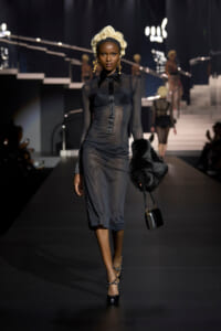 Model walks a dark runway wearing a sheer black midi dress with a fitted bodice and button-down front, black heels, and a furry black bag.