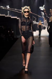 Model walks a fashion show runway in a black sheer mesh dress over lingerie, with oversized sunglasses, blonde curls, and a small black clutch.