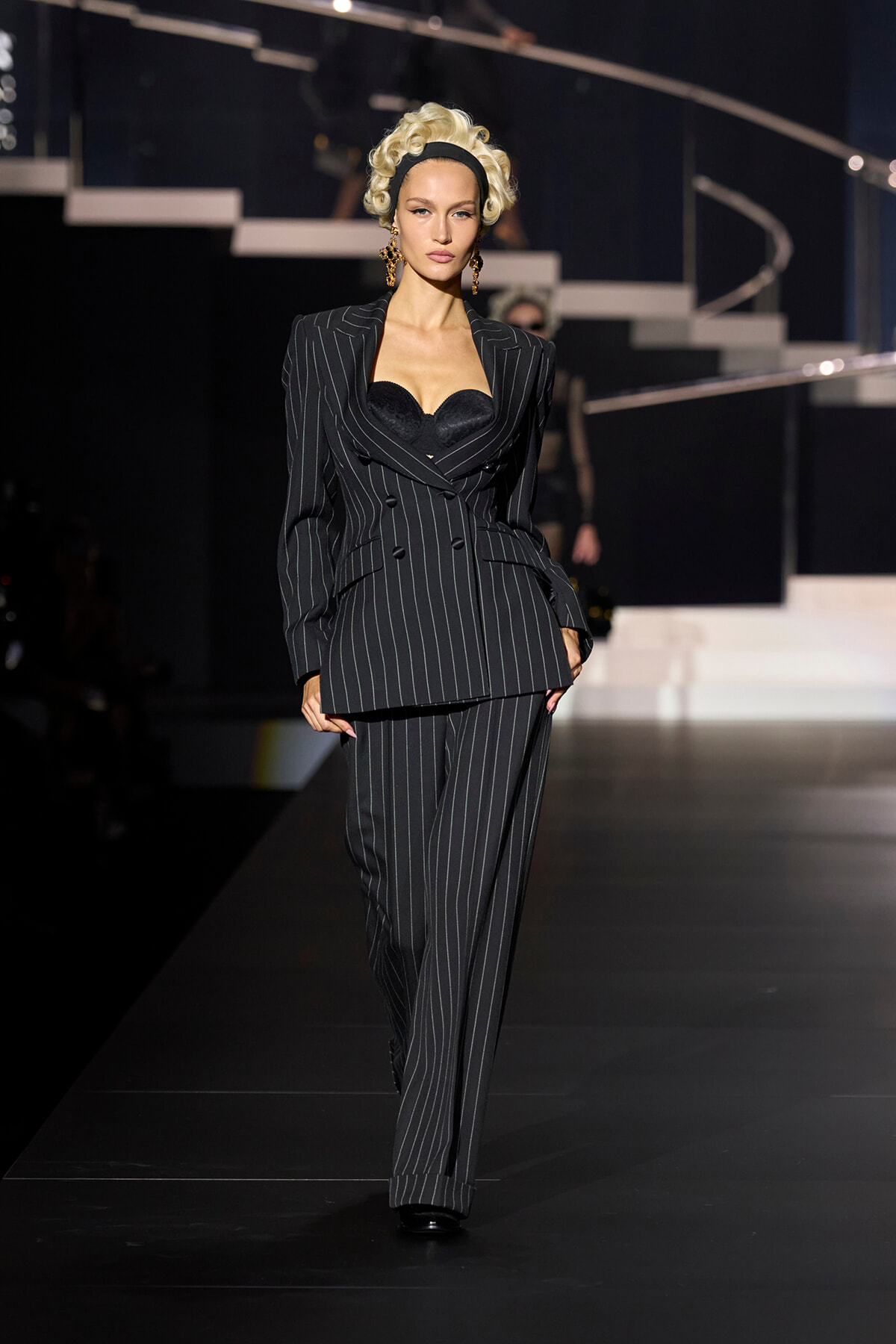 Model walking a runway in a black pinstripe double-breasted suit with a matching wide-leg trouser, revealing a black bustier top and gold earrings.