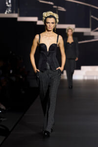Model on a runway wearing a black corset top and pinstriped trousers, blonde hair with a black headband, mid-walk pose.