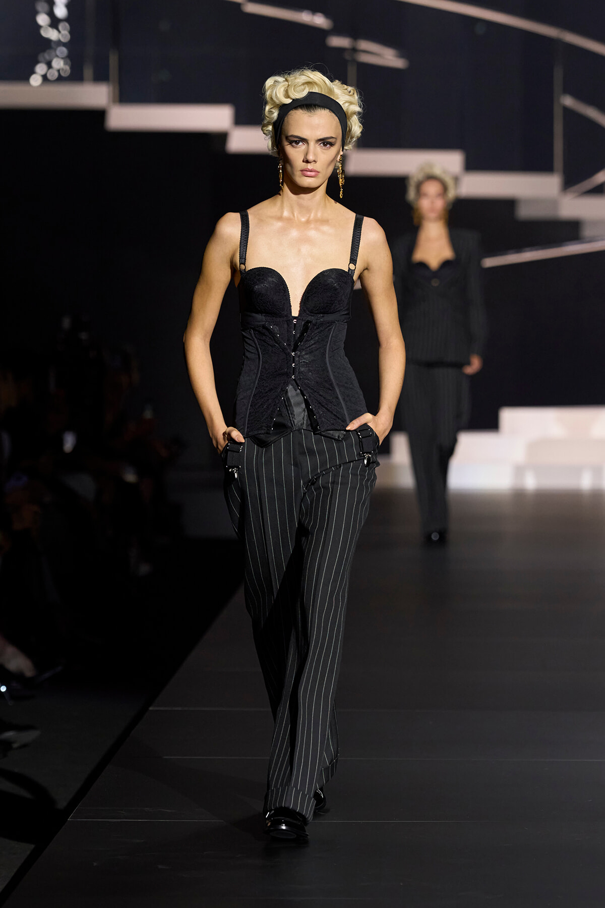 Model on a runway wearing a black corset top and pinstriped trousers, blonde hair with a black headband, mid-walk pose.